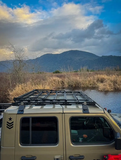 Jeep Gladiator with a roof rack parked near water and mountains under a cloudy sky.